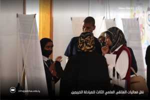 Group of young adults in hijabs and masks discuss near information boards at an indoor conference. The scene is foregrounded by poster boards and a few attendees engage in conversation.