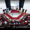 Audience seated in a large auditorium watching a panel discussion on stage with banners and a Libyan flag behind them.