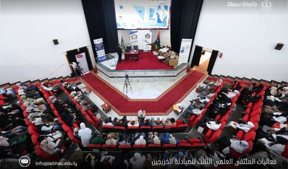 Audience seated in a large auditorium watching a panel discussion on stage with banners and a Libyan flag behind them.