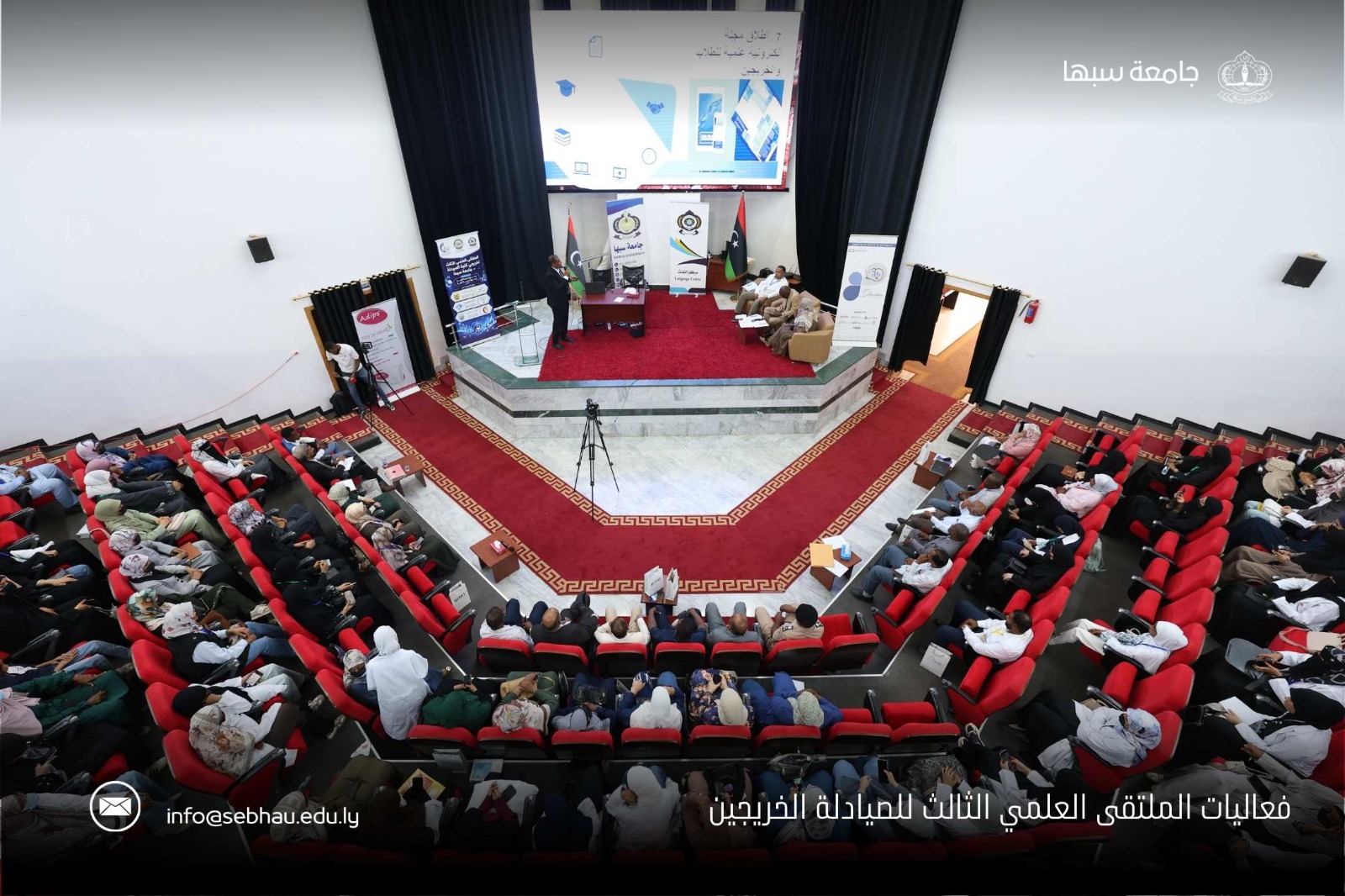 Audience seated in a large auditorium watching a panel discussion on stage with banners and a Libyan flag behind them.