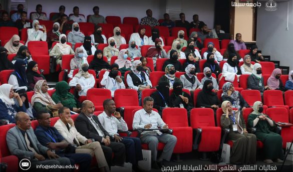 6EBE2E2F-6494-46C1-B88C-497E253AD6EF - جامعة سبها | Sebha University Audience seated in red theater chairs at a conference, many attendees wearing masks, front row with men in suits and women in traditional dress.