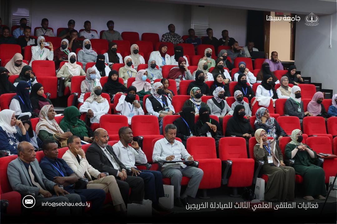 Audience seated in red theater chairs at a conference, many attendees wearing masks, front row with men in suits and women in traditional dress.