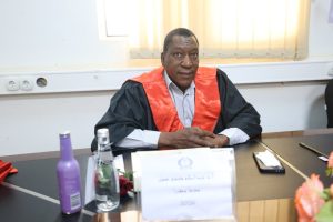 6F41E6B6-6054-47BE-AD00-2E591766CCB3 - جامعة سبها | Sebha University Man in black academic gown with a red sash sits at a conference table, hands folded, looking at the camera.