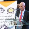 A man in a suit speaks at a glass podium during a conference, with Arabic banners and logos in the background. The banner shows organizational emblems and Arabic text behind him.