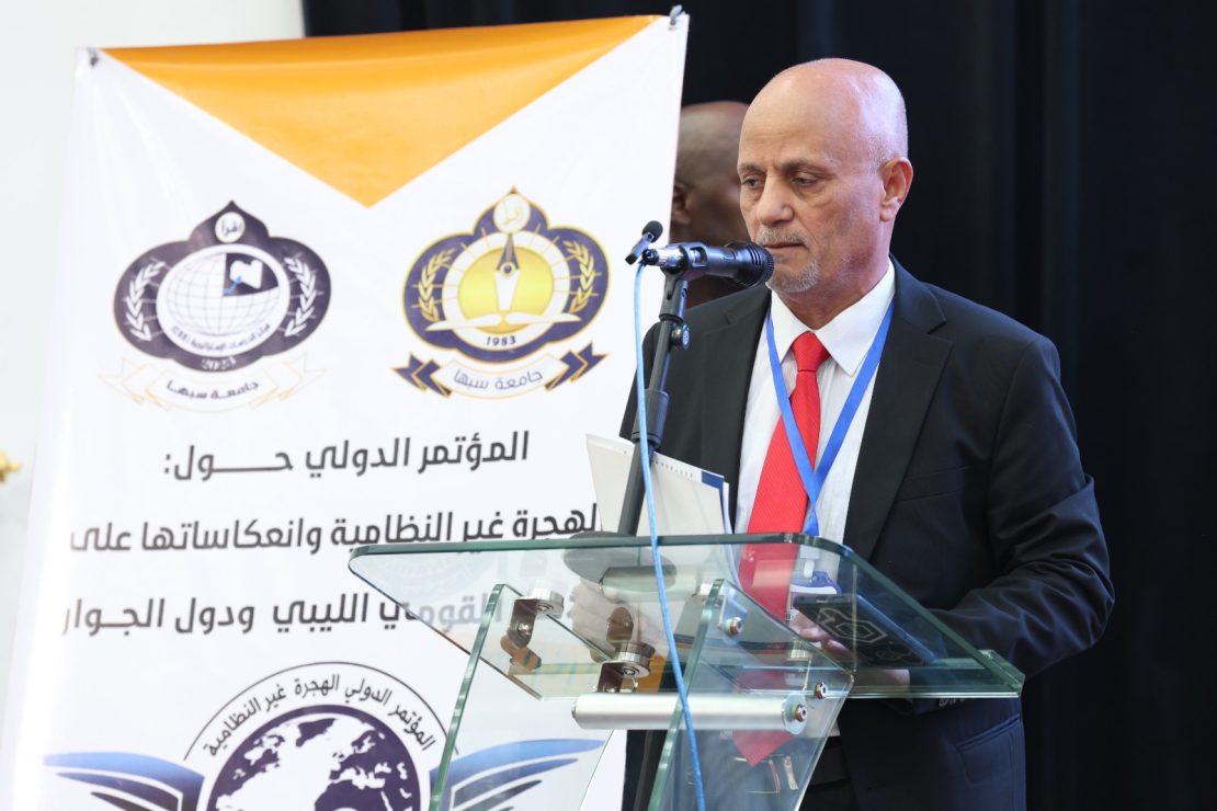 A man in a suit speaks at a glass podium during a conference, with Arabic banners and logos in the background. The banner shows organizational emblems and Arabic text behind him.
