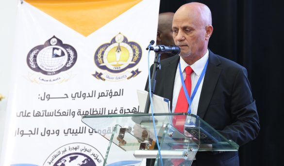 71A6A9B6-537A-4E98-93FE-0D2CF91B69B7 - جامعة سبها | Sebha University A man in a suit speaks at a glass podium during a conference, with Arabic banners and logos in the background. The banner shows organizational emblems and Arabic text behind him.