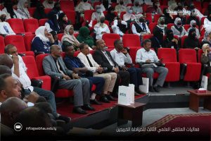 Audience seated in red theater seats at a conference, diverse attendees including women in hijabs; conference bags on the floor nearby.