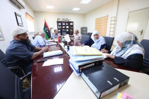 People gathered around a long conference table reviewing documents, with flags and a cabinet in the background of a formal meeting room.