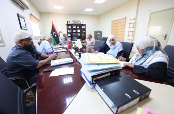 People gathered around a long conference table reviewing documents, with flags and a cabinet in the background of a formal meeting room.