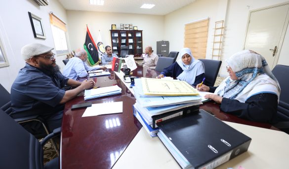 People gathered around a long conference table reviewing documents, with flags and a cabinet in the background of a formal meeting room.