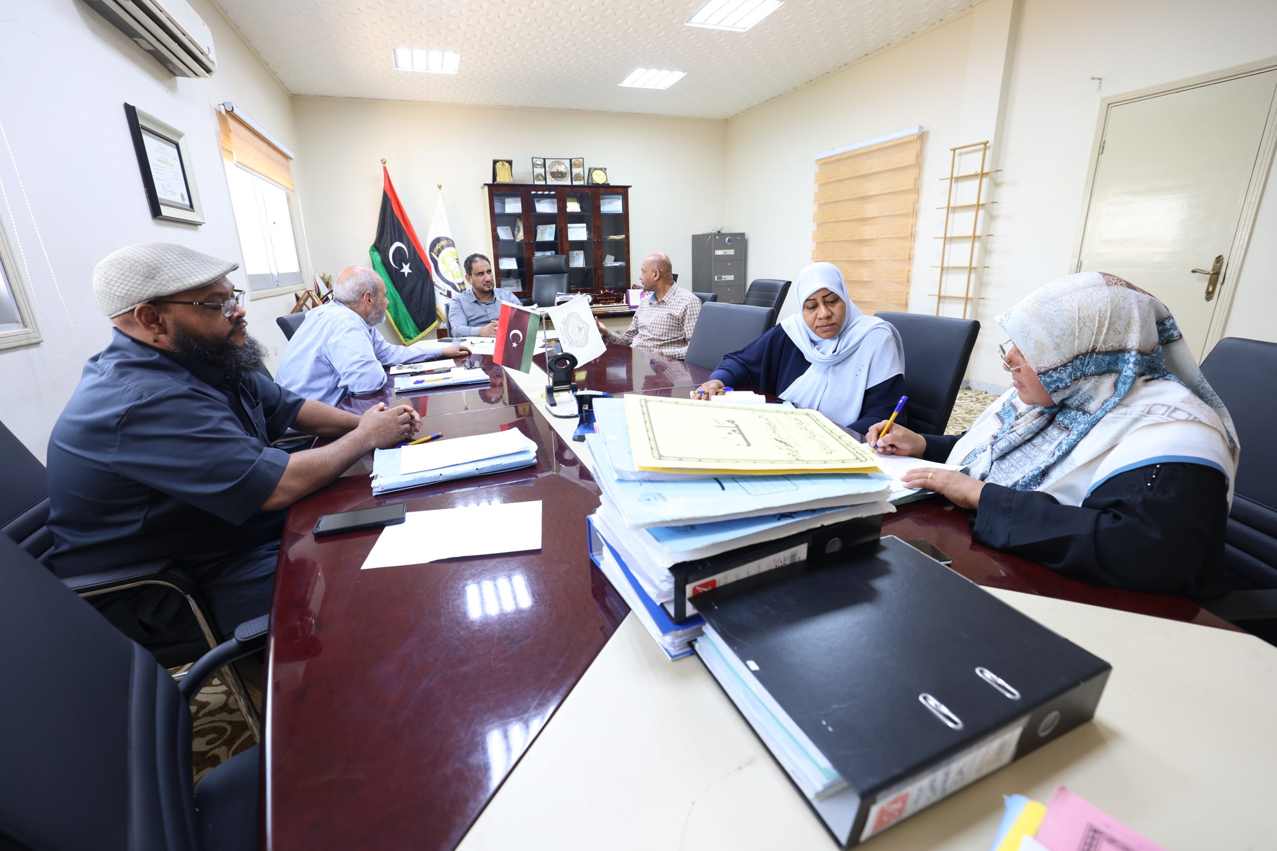 People gathered around a long conference table reviewing documents, with flags and a cabinet in the background of a formal meeting room.