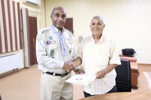 Two men shake hands in an office while one hands over a folder; one wears a badge-filled uniform, the other a striped shirt.