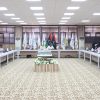 Formal meeting in a conference room with participants seated along a U-shaped table and flags displayed behind the speakers.