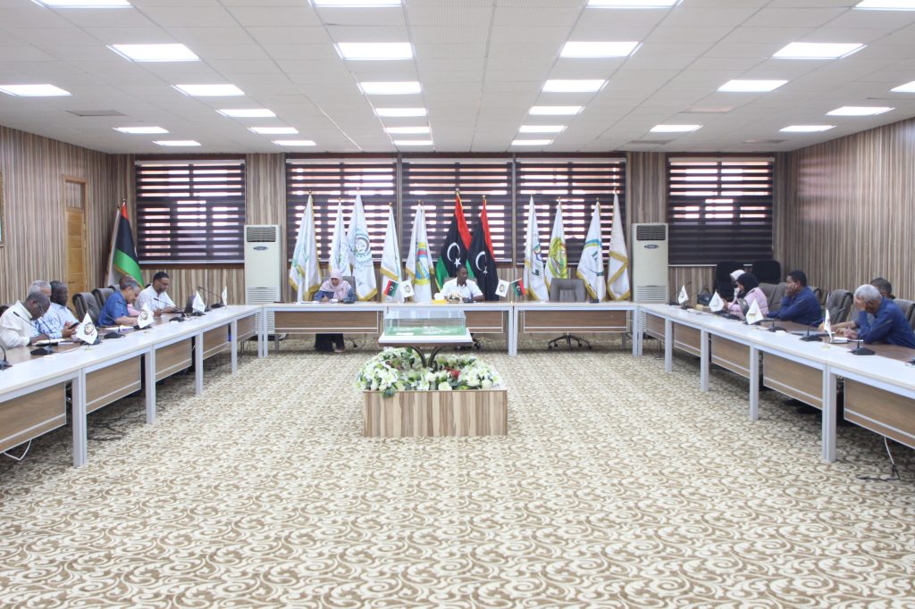 Formal meeting in a conference room with participants seated along a U-shaped table and flags displayed behind the speakers.