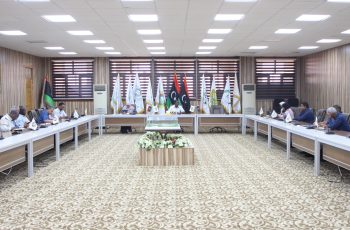 Formal meeting in a conference room with participants seated along a U-shaped table and flags displayed behind the speakers.