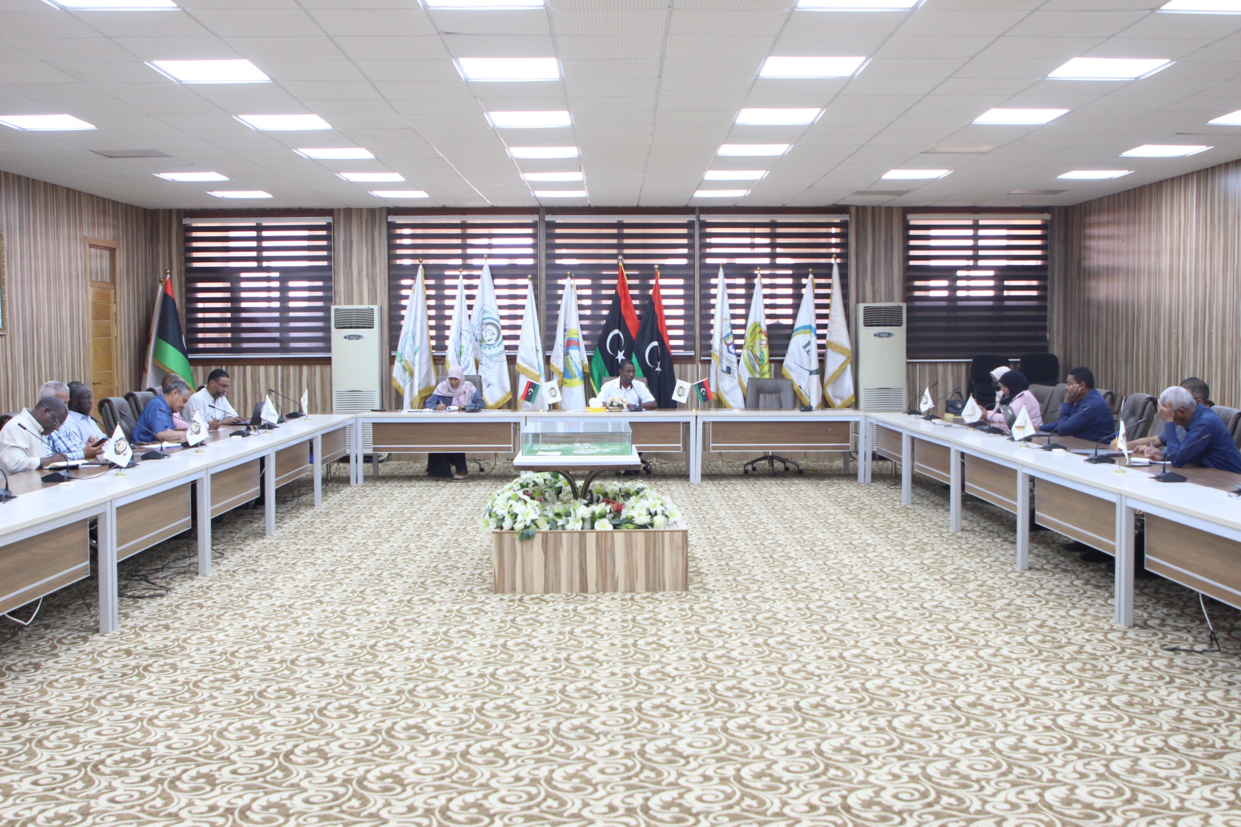 Formal meeting in a conference room with participants seated along a U-shaped table and flags displayed behind the speakers.