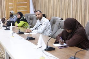 Man in a patterned white shirt sits at a conference table, looking at the camera, with others in hijabs beside him and microphones on the table behind a wood-paneled wall.