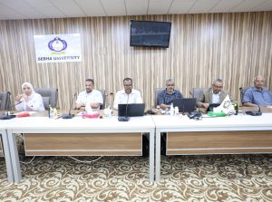 Six professionals seated at a long conference table in a meeting room, with a Sebha University banner on a wood-paneled wall behind them.