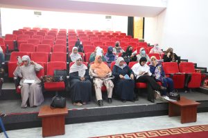 Audience of women wearing hijabs seated in a red-upholstered auditorium; several hold papers or phones and wear masks.