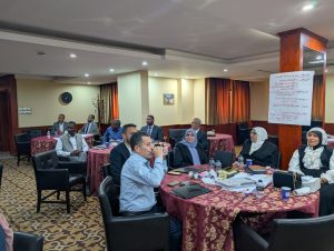Group of professionals seated around round tables in a hotel conference room, some taking notes and talking, with a poster on a pillar nearby.