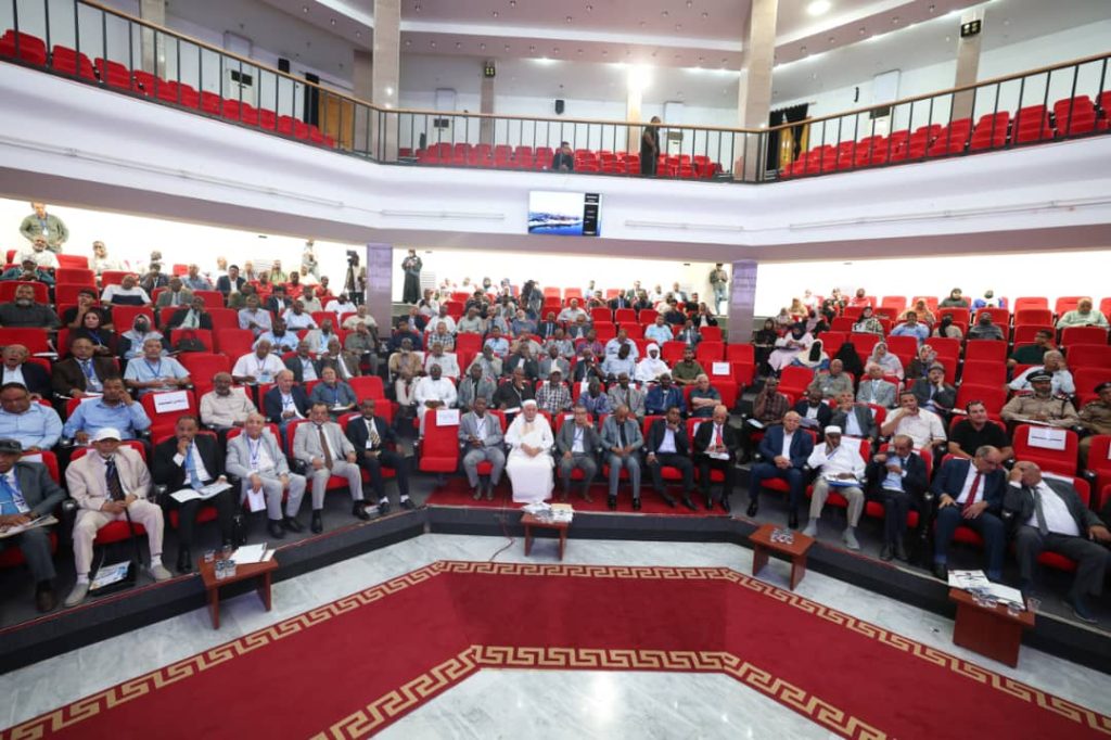 Wide view of a formal conference hall filled with attendees seated in red chairs on multiple tiers.