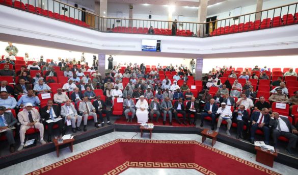 Wide view of a formal conference hall filled with attendees seated in red chairs on multiple tiers.