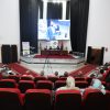 Audience seated in red theater chairs watching a panel on a stage with a large screen showing a speaker and banners behind them on a podium area in a modern auditorium.