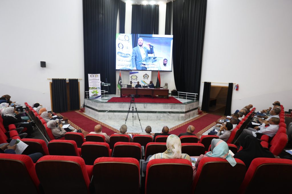 86AF6775-041F-419E-8C70-30E8CC9F1F0C - جامعة سبها | Sebha University Audience seated in red theater chairs watching a panel on a stage with a large screen showing a speaker and banners behind them on a podium area in a modern auditorium.
