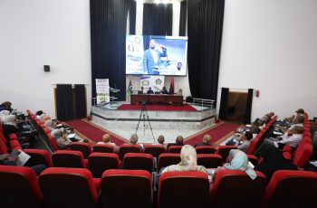 Audience seated in red theater chairs watching a panel on a stage with a large screen showing a speaker and banners behind them on a podium area in a modern auditorium.