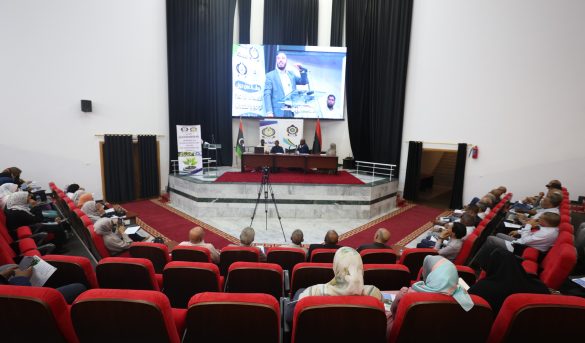 86AF6775-041F-419E-8C70-30E8CC9F1F0C - جامعة سبها | Sebha University Audience seated in red theater chairs watching a panel on a stage with a large screen showing a speaker and banners behind them on a podium area in a modern auditorium.