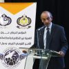 Speaker in a dark suit at a glass podium delivering a speech beside a white banner with logos and Arabic text in the background.