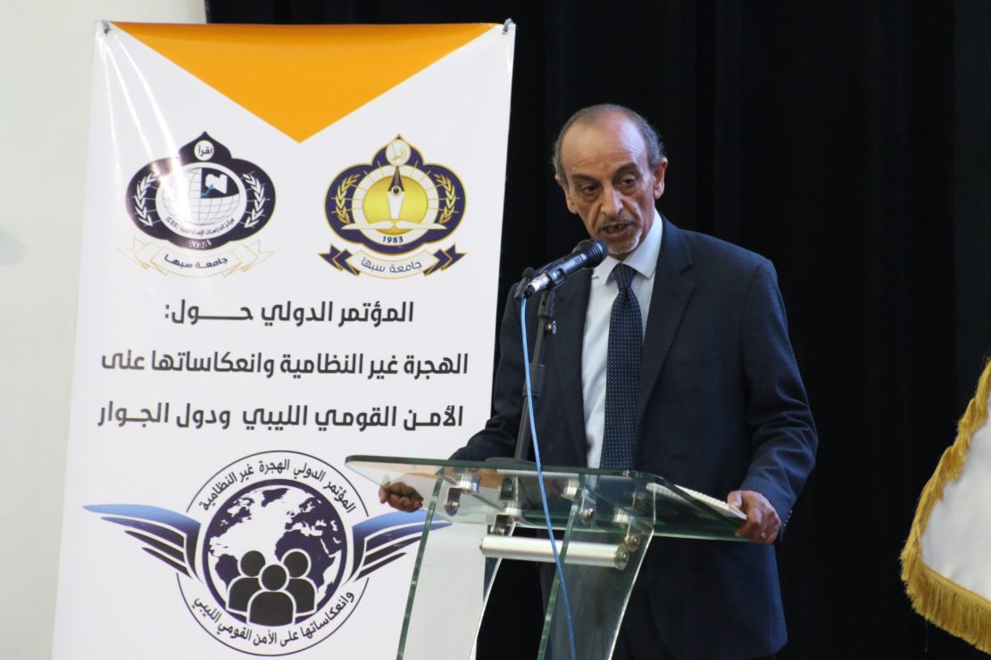 Speaker in a dark suit at a glass podium delivering a speech beside a white banner with logos and Arabic text in the background.