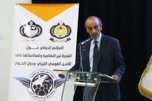 89318A1F-0175-413A-8C97-277BD249F809 - جامعة سبها | Sebha University Speaker in a dark suit at a glass podium delivering a speech beside a white banner with logos and Arabic text in the background.