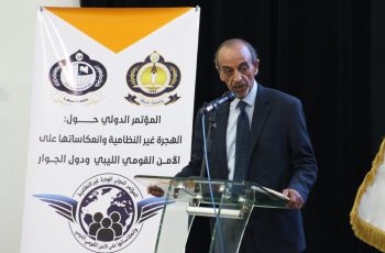 Speaker in a dark suit at a glass podium delivering a speech beside a white banner with logos and Arabic text in the background.