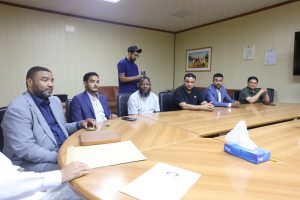 89CD78B3-F827-4AC1-AE4C-3018B302CF5C - جامعة سبها | Sebha University Group of men seated around a conference table in a meeting room, one man stands with a phone as if recording a selfie video of the group.