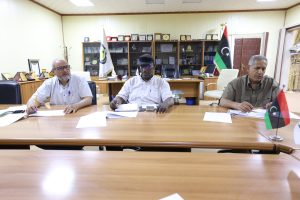 Three men sit at a long conference table signing papers in a trophy-filled office; a Libyan flag is visible in the background.