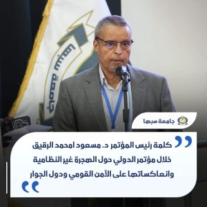 Man in a gray suit and glasses speaks into a microphone at a podium on a stage, with a flag in the background and a large white caption panel containing Arabic text at the bottom right.