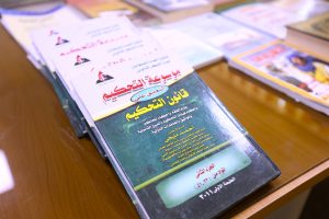 Stack of Arabic-language textbooks on a wooden table; the foreground book has a green cover with yellow Arabic text.