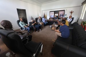 Group of men seated in a meeting room around a central desk, engaged in discussion, with flags and framed portraits on the wall behind them.
