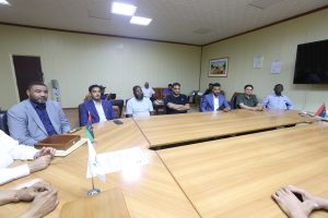 8FC38DAE-216F-416E-BA04-2C58197F9045 - جامعة سبها | Sebha University Group of men seated along a long conference table in a beige-walled meeting room, with small flags on the table and a large screen on the wall.