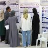 Group of people in traditional clothing read large posters with Arabic text and diagrams on a wall at an indoor exhibition or conference.