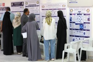 Group of people in traditional clothing read large posters with Arabic text and diagrams on a wall at an indoor exhibition or conference.