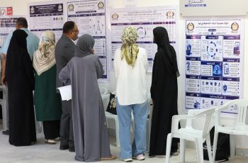 Group of people in traditional clothing read large posters with Arabic text and diagrams on a wall at an indoor exhibition or conference.