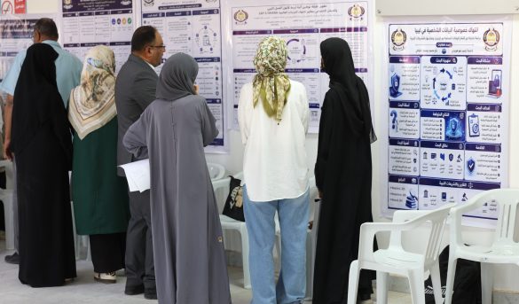90533C4C-DCC2-4882-BE14-05F4C5268C2F - جامعة سبها | Sebha University Group of people in traditional clothing read large posters with Arabic text and diagrams on a wall at an indoor exhibition or conference.