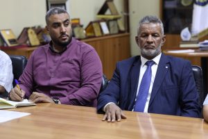938CF9E1-AB21-497B-B630-6A22E6DBCF4A - جامعة سبها | Sebha University Two men seated at a wooden conference table in an office; the man on the left writes in a notebook, the man on the right wears a blue suit and looks ahead.