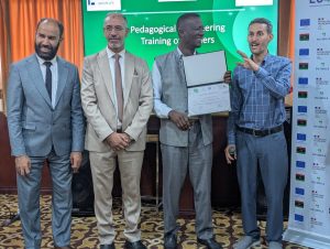 9A167D43-A080-4F26-A3D9-9DDCDF897621 - جامعة سبها | Sebha University Four men in business suits posing on stage at an award ceremony, one holding a certificate with a smile in the background banner behind them.