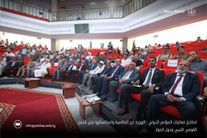 Audience seated in red-tiered auditorium for a formal international conference, with officials in front rows and a university backdrop (Sebha University logo visible).