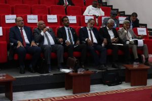 9D3CEFB2-DDBF-4E00-A69C-169B22BB19BC - جامعة سبها | Sebha University Row of men in business suits seated in red auditorium chairs during a conference, some with papers and briefcases nearby.