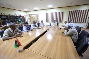 Officials sit around a long conference table in a meeting room, with a Libyan flag on the table and shelves in the background.