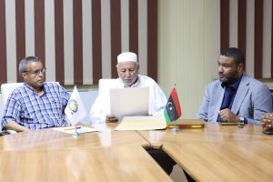 9DC59756-4CAC-4C68-84E3-758A5A22CA73 - جامعة سبها | Sebha University Three men seated at a conference table looking at documents, with flags on the table in front of them.